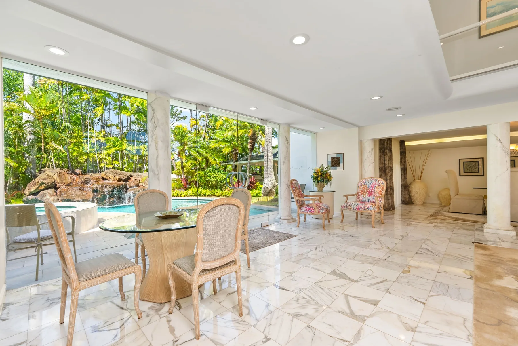 Una luminosa sala de estar con una mesa junto a grandes ventanales que dan al jardín y la piscina.