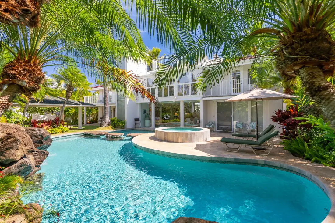 A swimming pool curves around a raised circular spa beside a glass-fronted house.