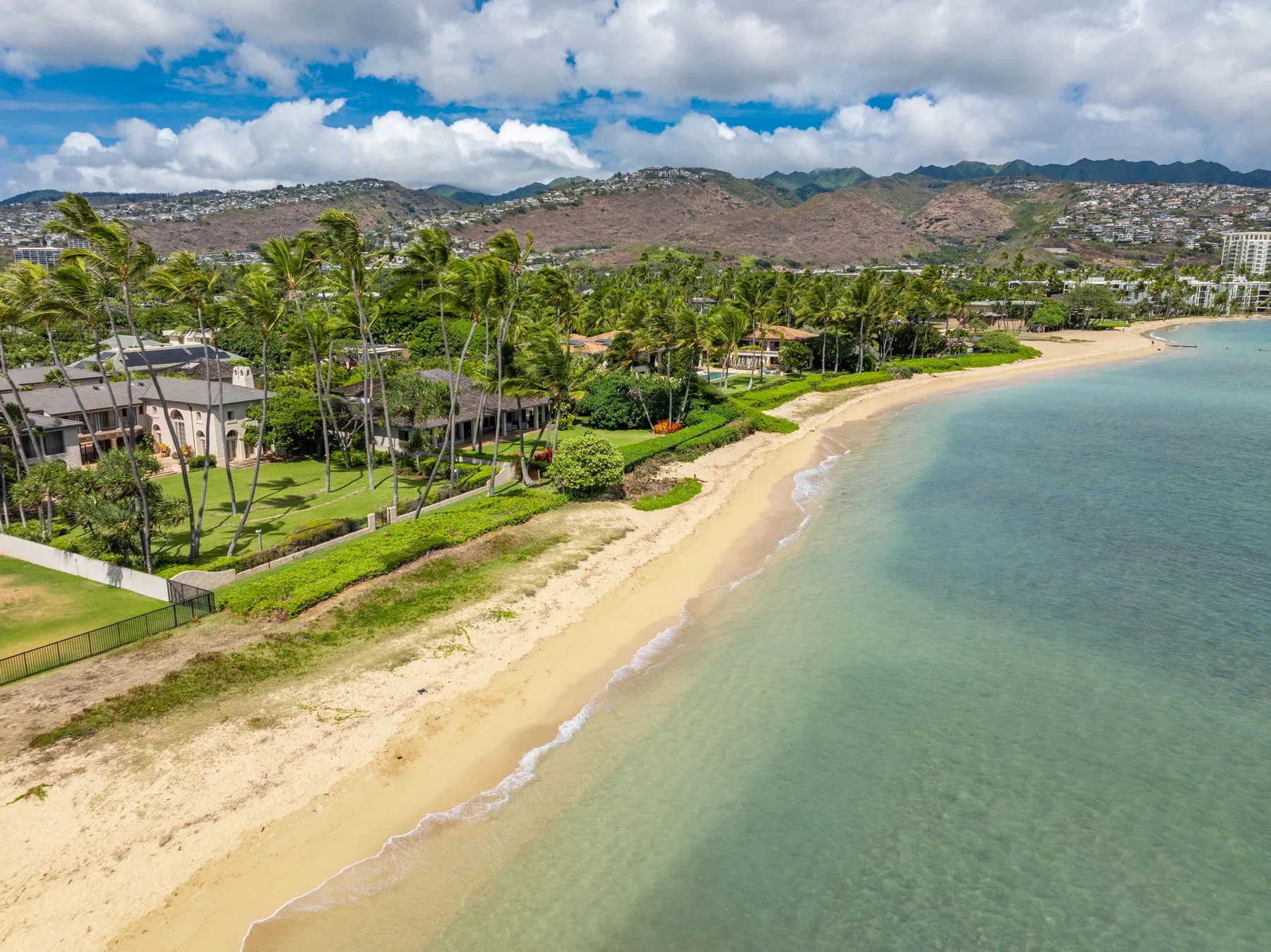 Una playa de arena bordea la costa junto a palmeras y zonas verdes.