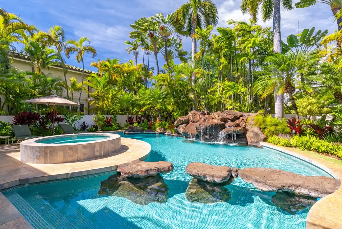 Una piscina tropical con un spa circular elevado y una cascada de roca.