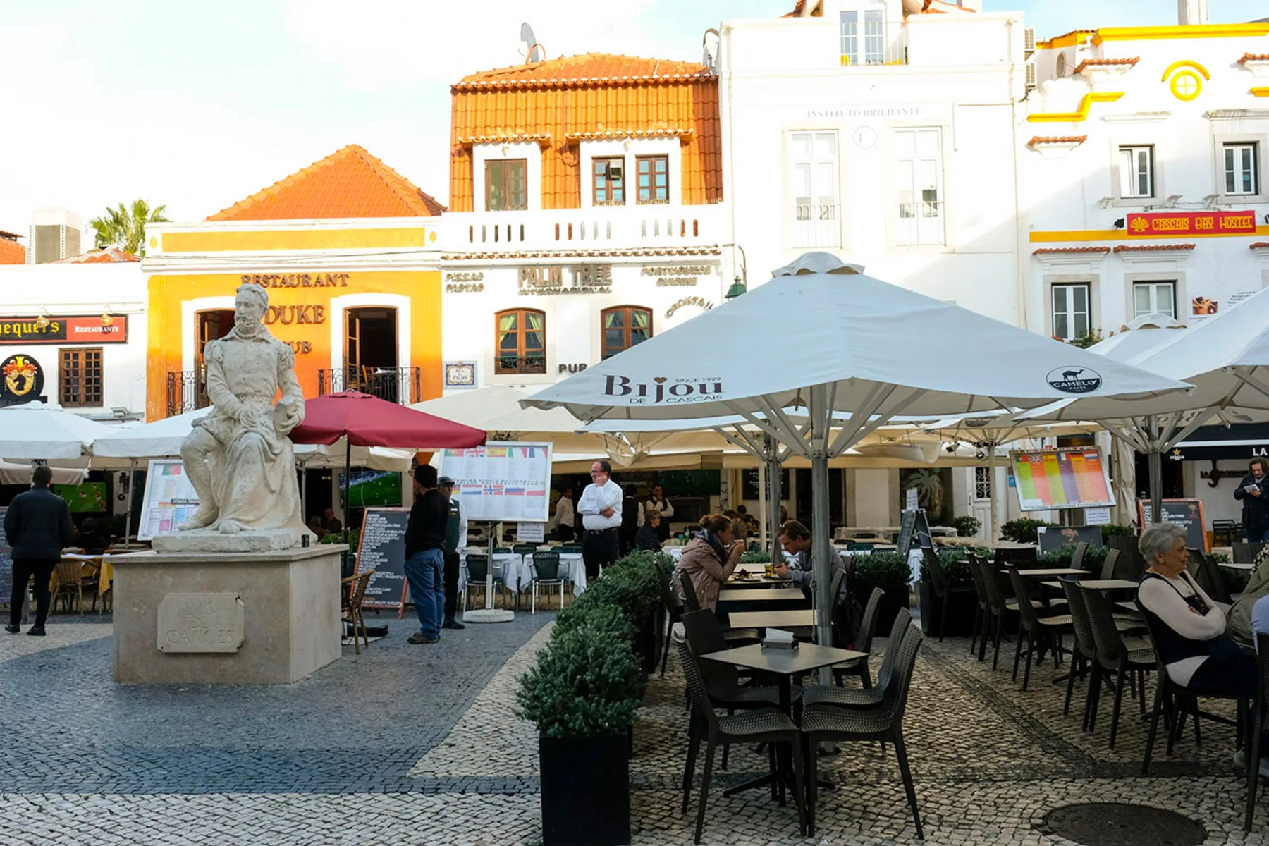 Una plaza con cafetería al aire libre, mesas a la sombra y una estatua de piedra.