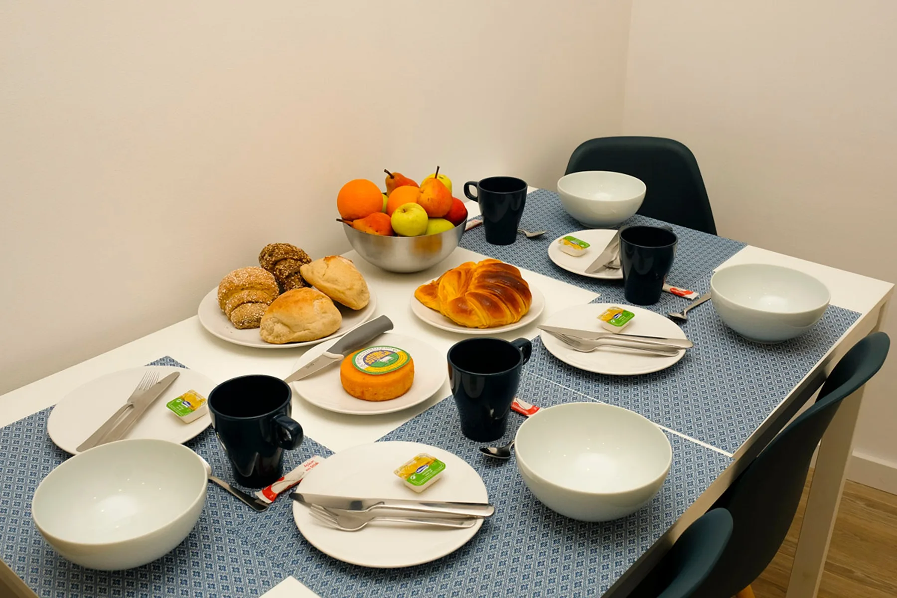 Una mesa de comedor está puesta con platos, tazas, pan y fruta.