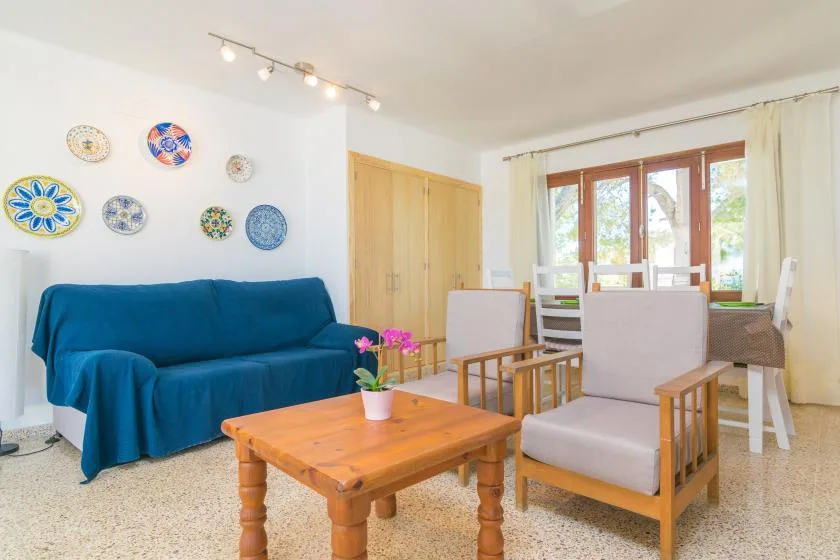 A bright living room with blue seating, a wooden coffee table, and a dining area by the windows.