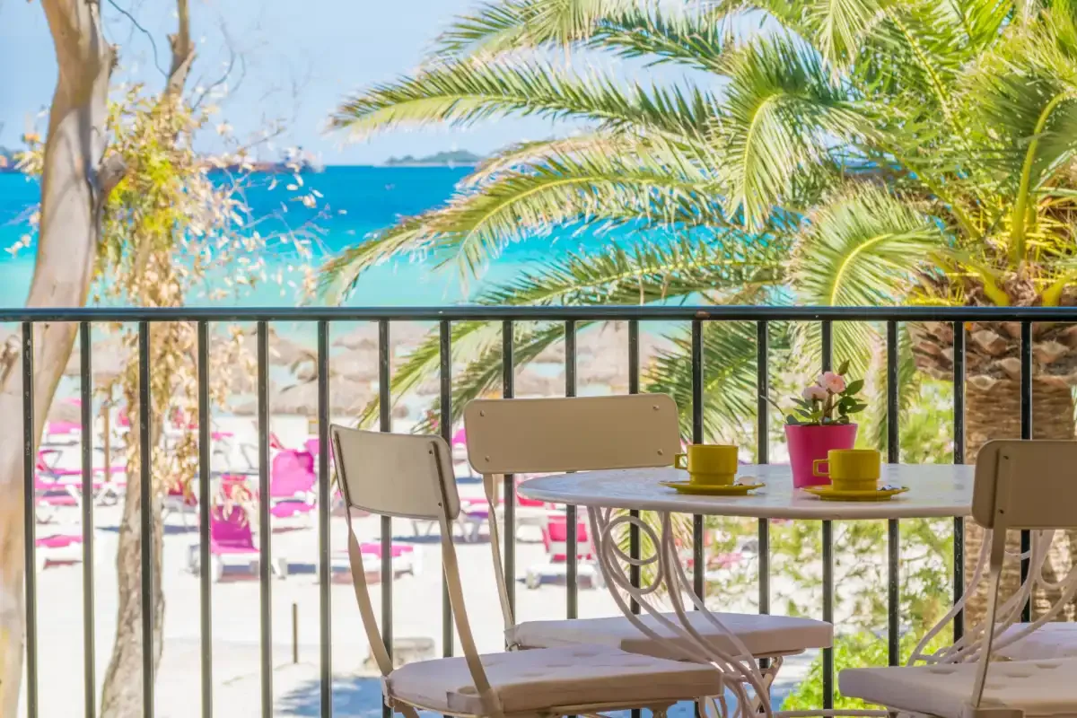 A balcony dining area overlooks a beach with turquoise water and palm trees.