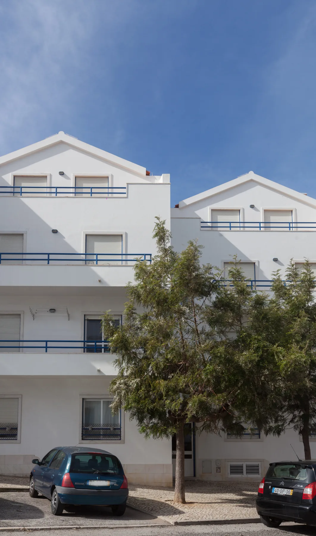 A white apartment building with balconies stands behind parked cars and trees.
