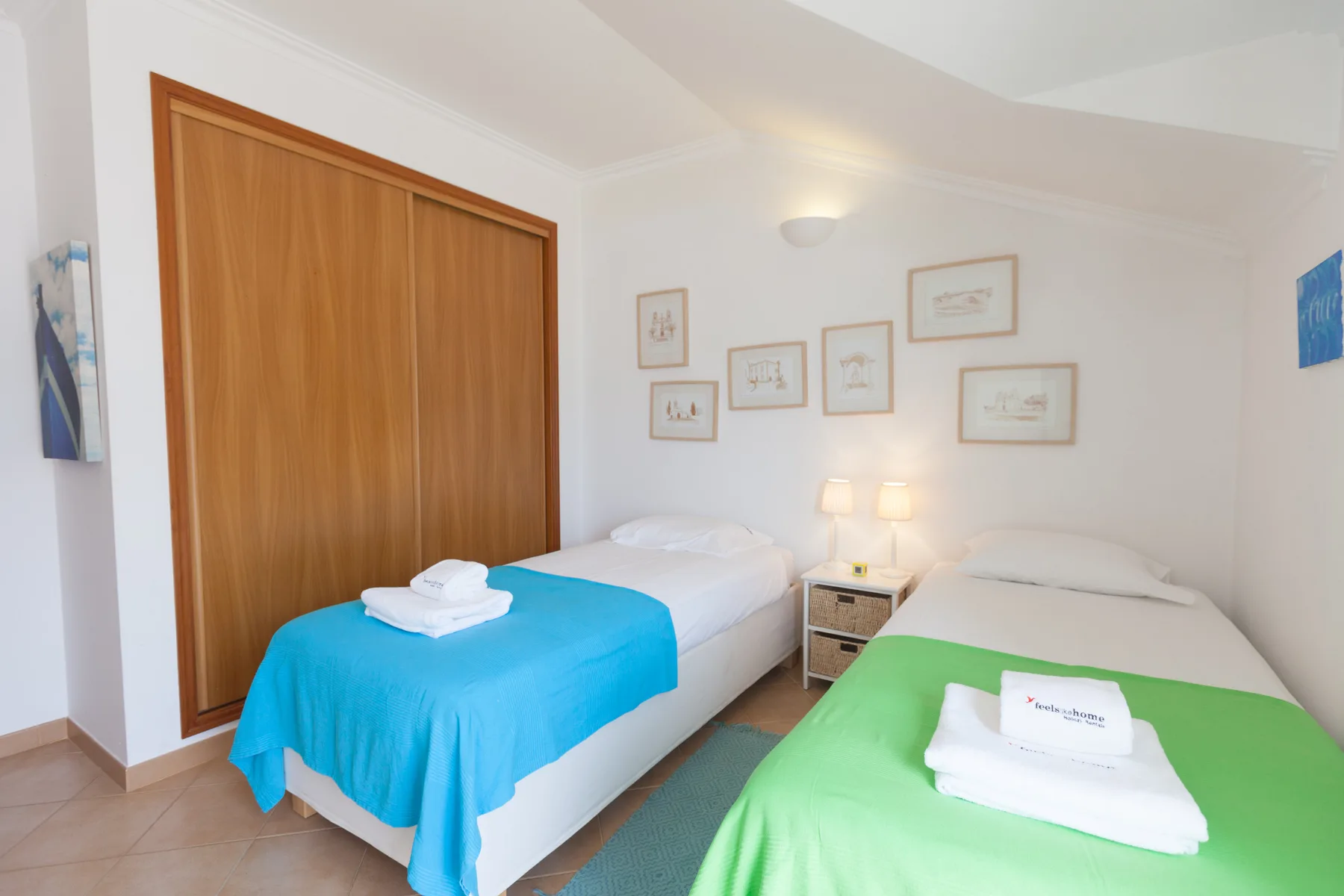 A bright bedroom with two beds, colorful bedspreads, and framed wall art.