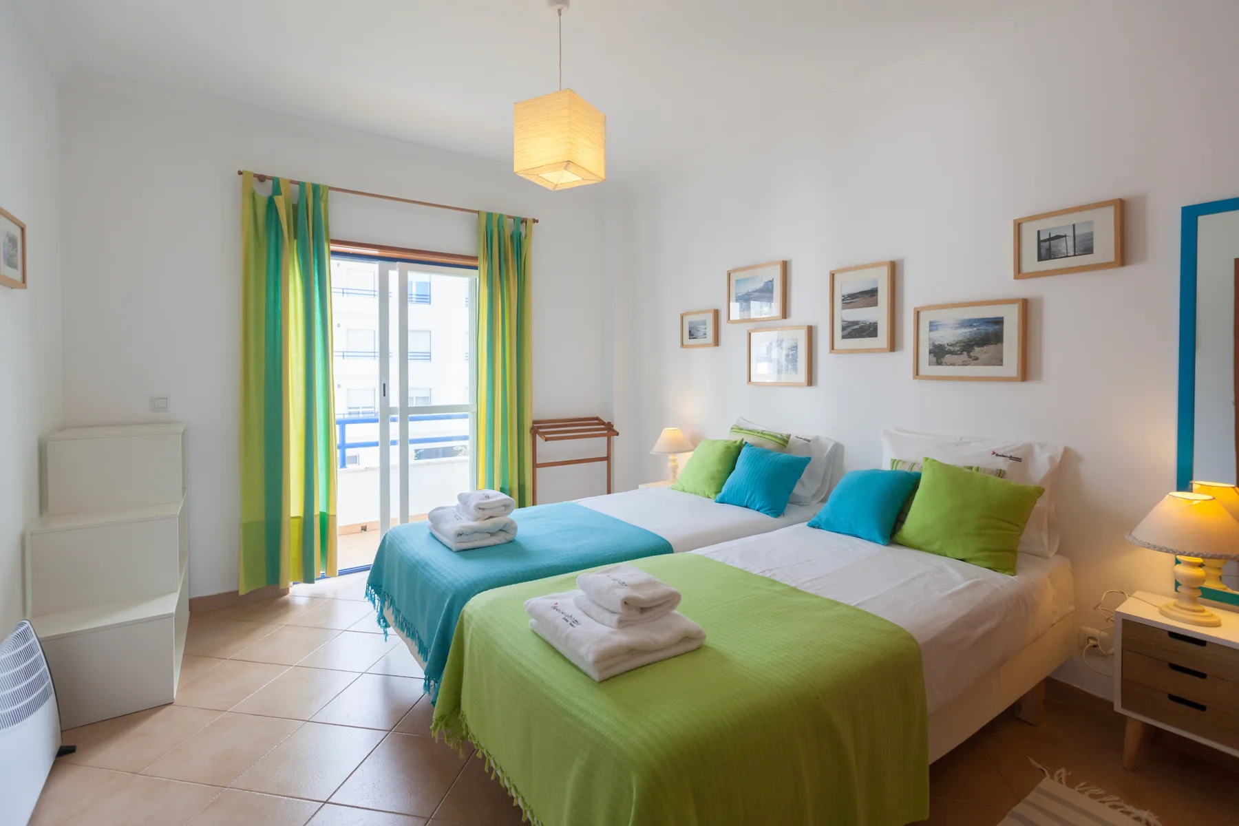 Un dormitorio luminoso con dos camas, ropa de cama colorida y una ventana con cortinas verdes.
