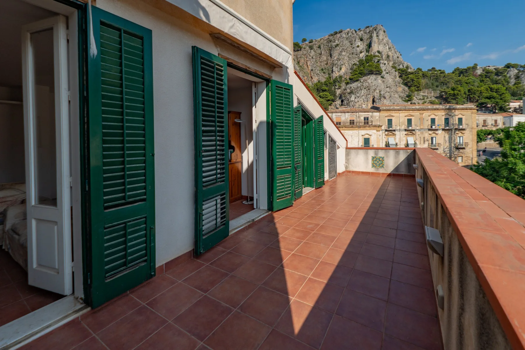 A tiled outdoor terrace with open doors and green shutters overlooks nearby buildings and rocky hills.