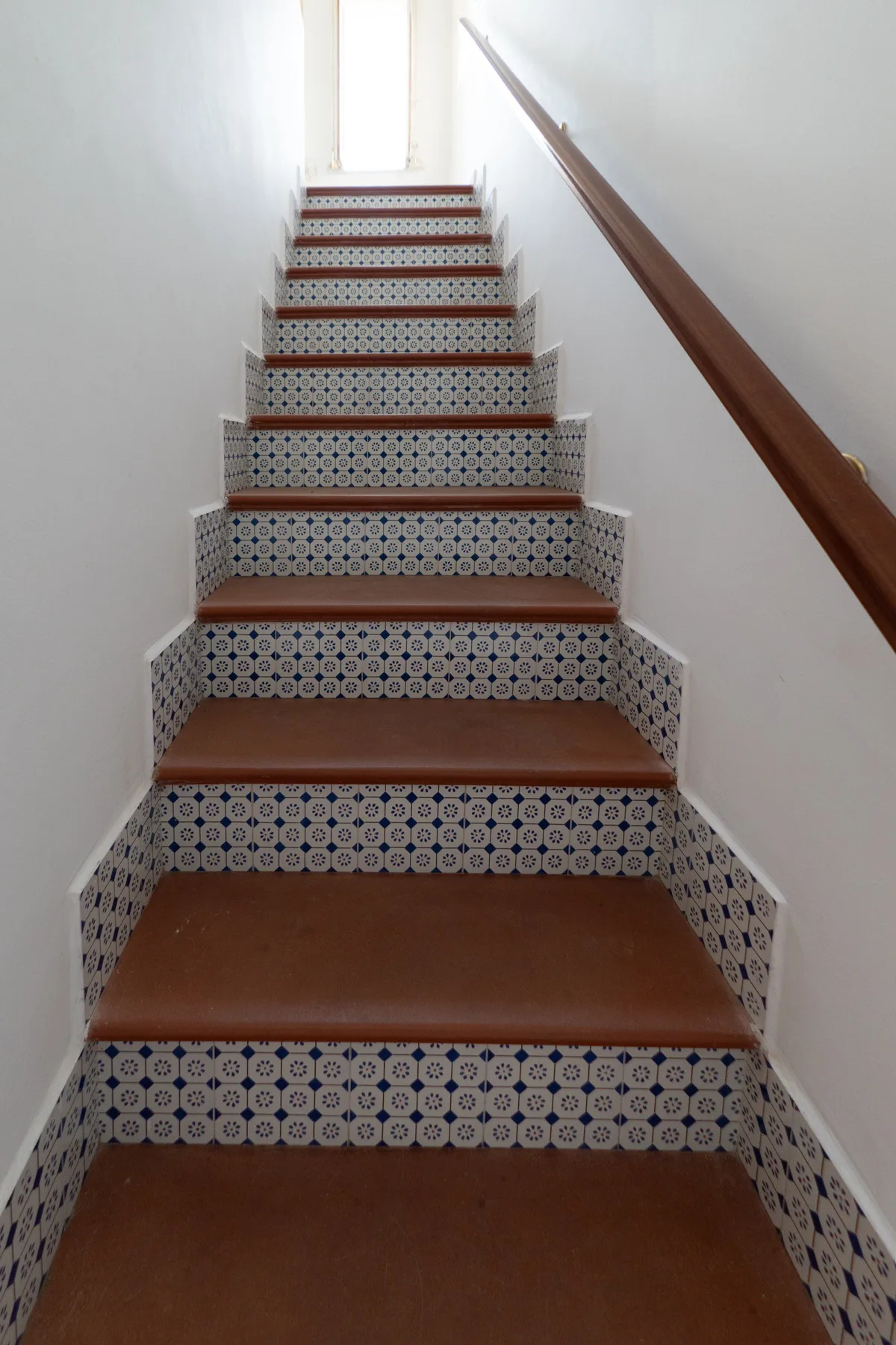 Una escalera estrecha con contrahuellas de azulejos estampados y un pasamanos de madera.