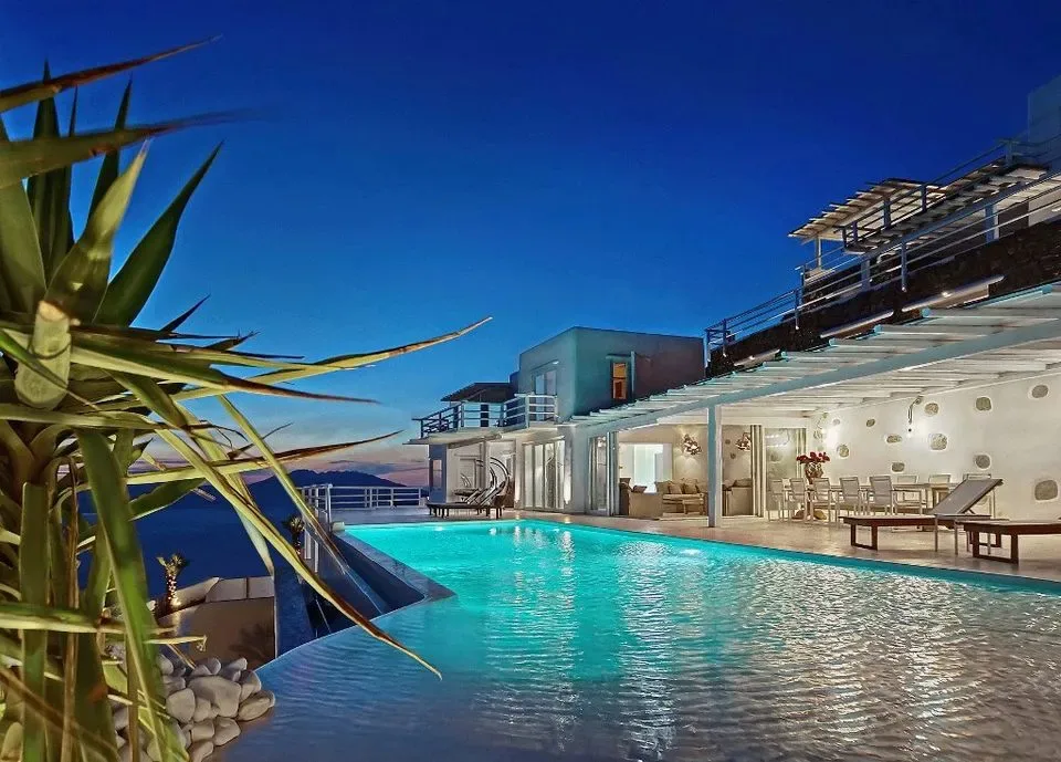 Un edificio moderno da a una piscina iluminada junto al mar al anochecer.
