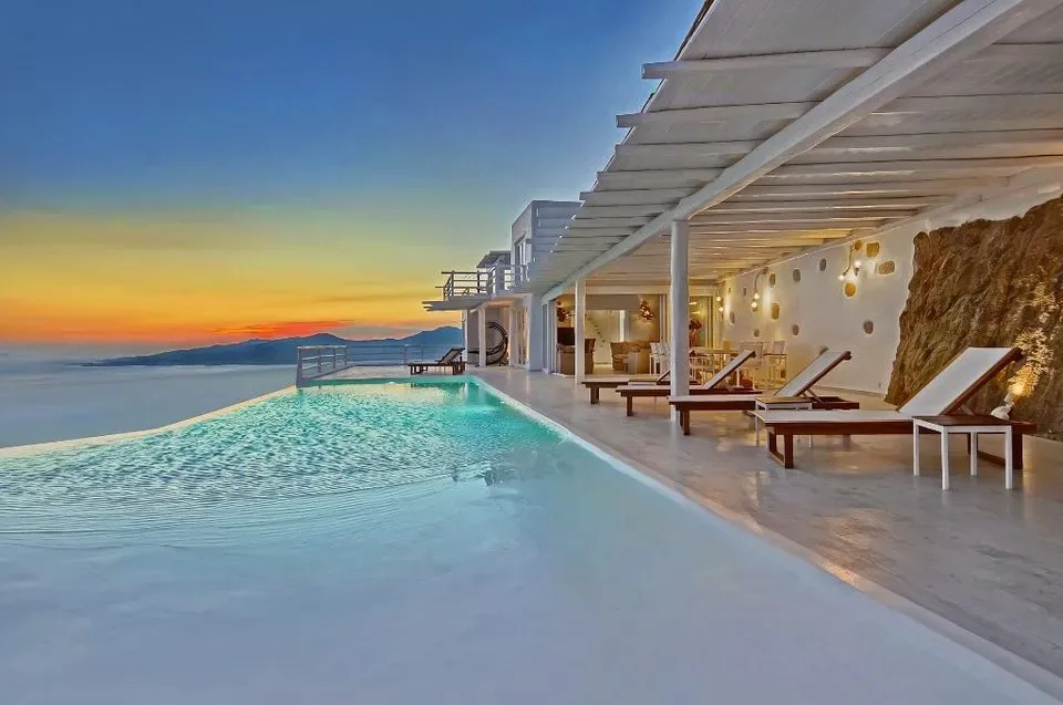 An infinity pool beside a covered terrace with seating overlooks the sea at sunset.