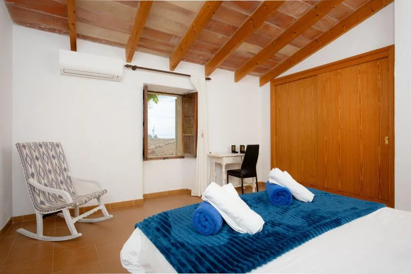 A bedroom with a blue bedspread, a rocking chair, and exposed wooden ceiling beams.