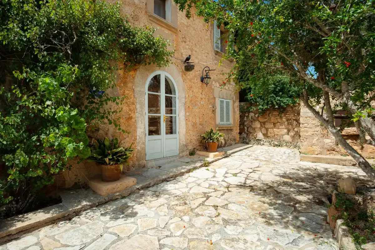 A stone-paved courtyard beside a rustic building, with greenery and a pale arched door.