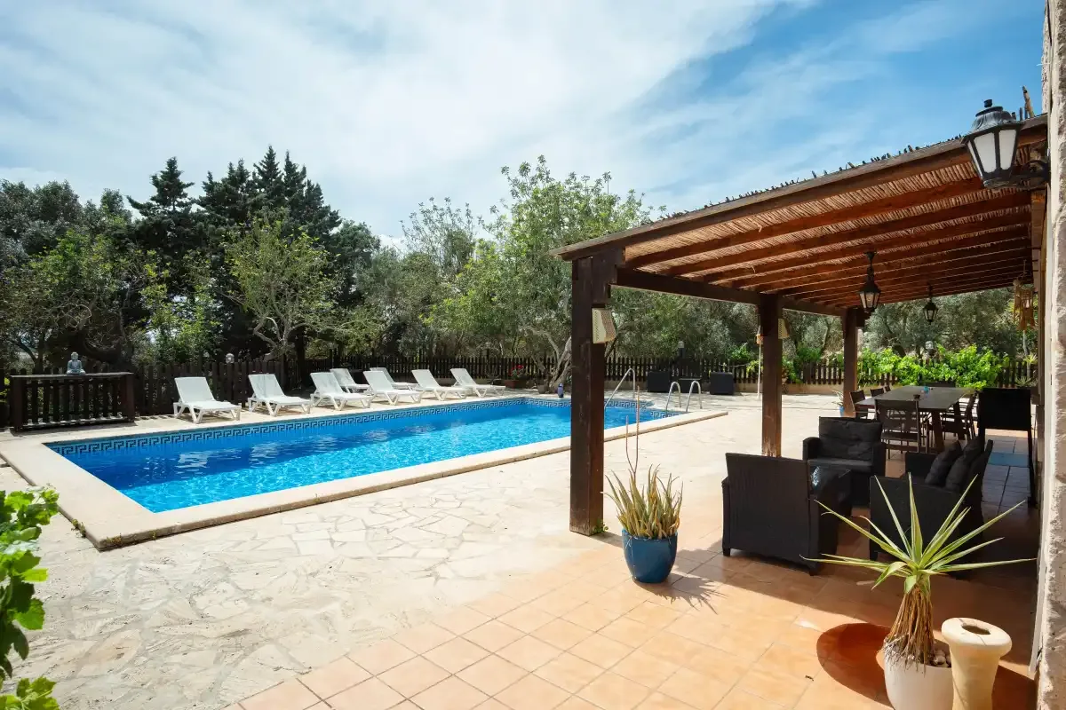 An outdoor pool beside a shaded patio with seating and surrounding trees.