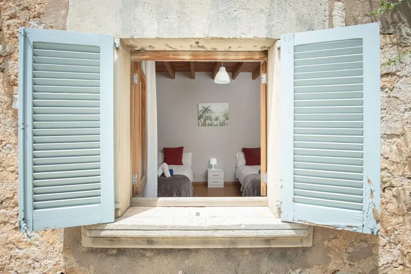 An open window looks into a bedroom with two beds and a small bedside table.