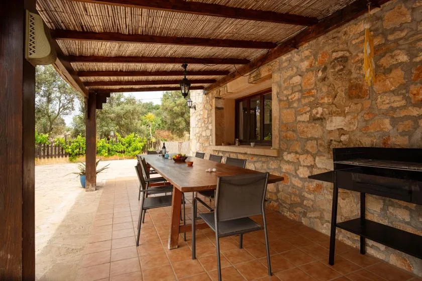 A covered stone patio with a long dining table and seating.