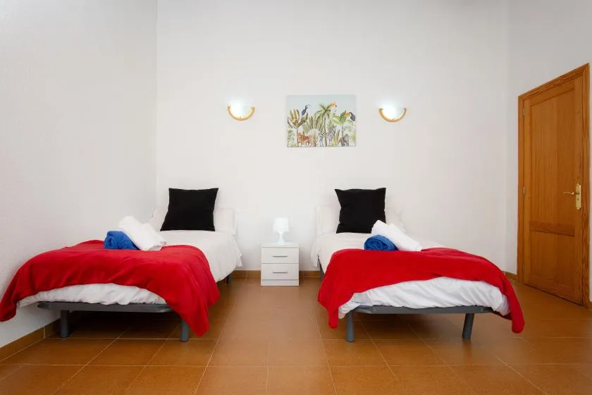 A bright bedroom with two beds, red bedspreads, and a small bedside table.