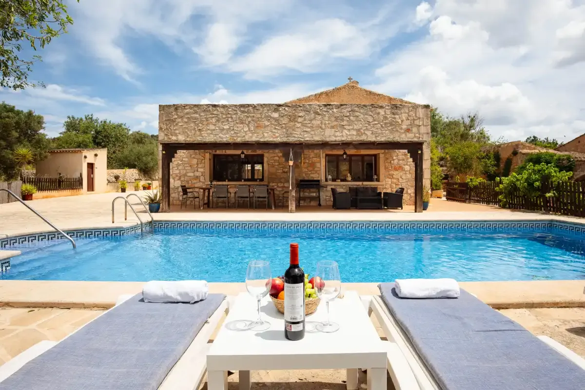 A stone house stands behind a swimming pool with loungers in the foreground.