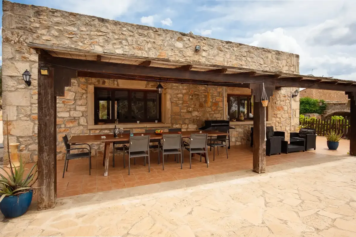 A covered stone patio has a dining table and seating beside the house.