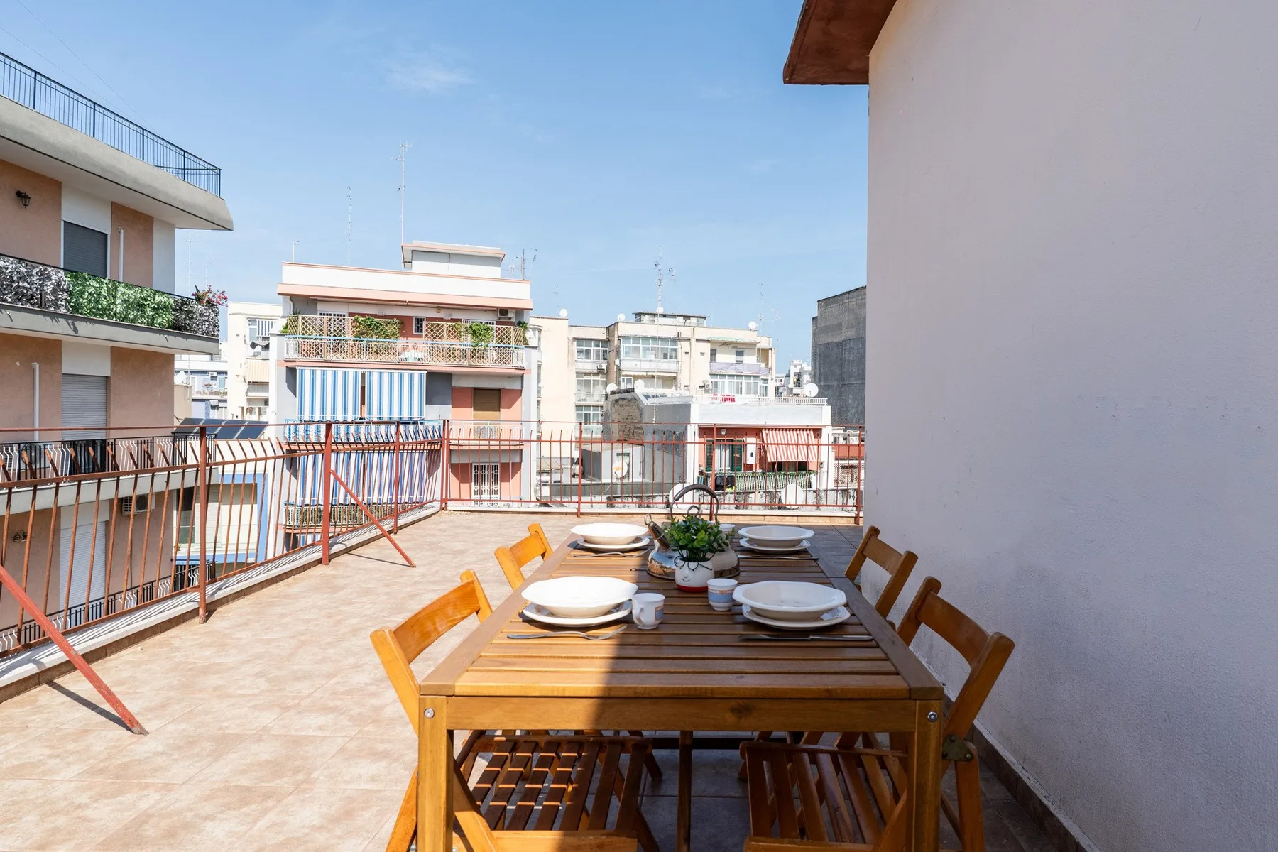 Una terraza en la azotea tiene una mesa de comedor de madera con platos y vistas a edificios cercanos.