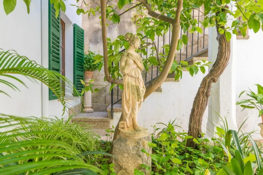 A leafy garden courtyard surrounds a classical statue beside a white building.