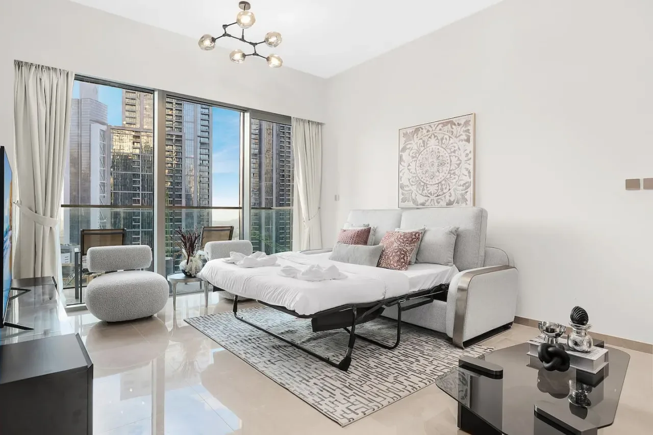 Un dormitorio luminoso con una cama junto a grandes ventanas y edificios a la vista.