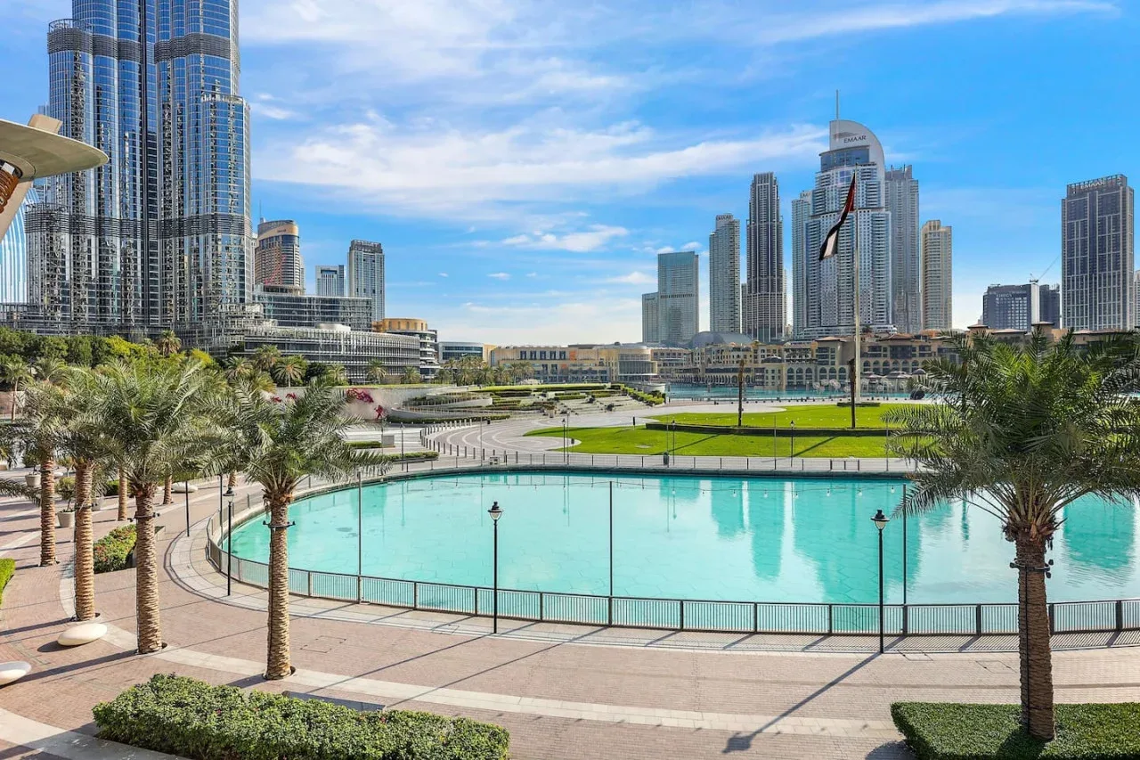 Una piscina turquesa y un paseo bordeado de palmeras se extienden ante un moderno horizonte urbano.