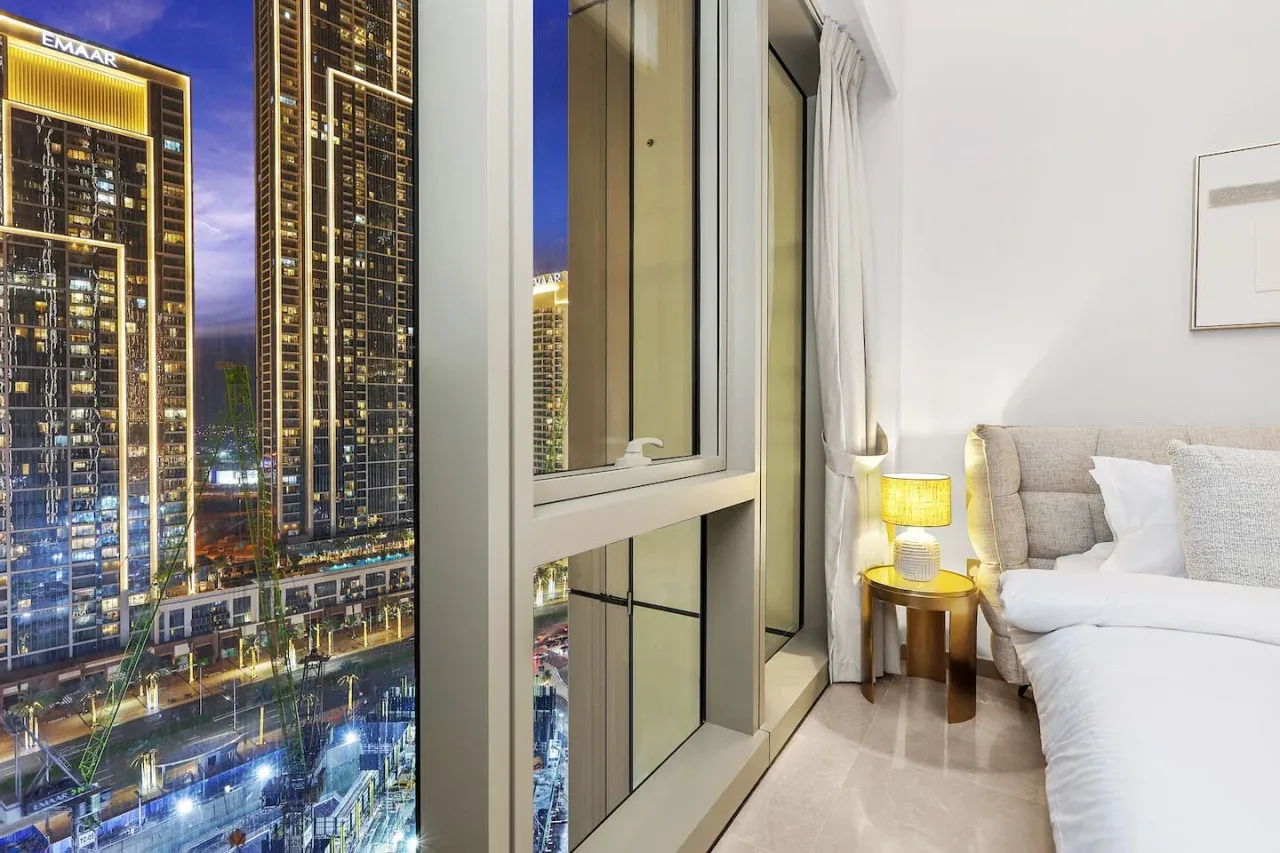 Dormitorio con una cama junto a una gran ventana con vistas a edificios iluminados por la noche.