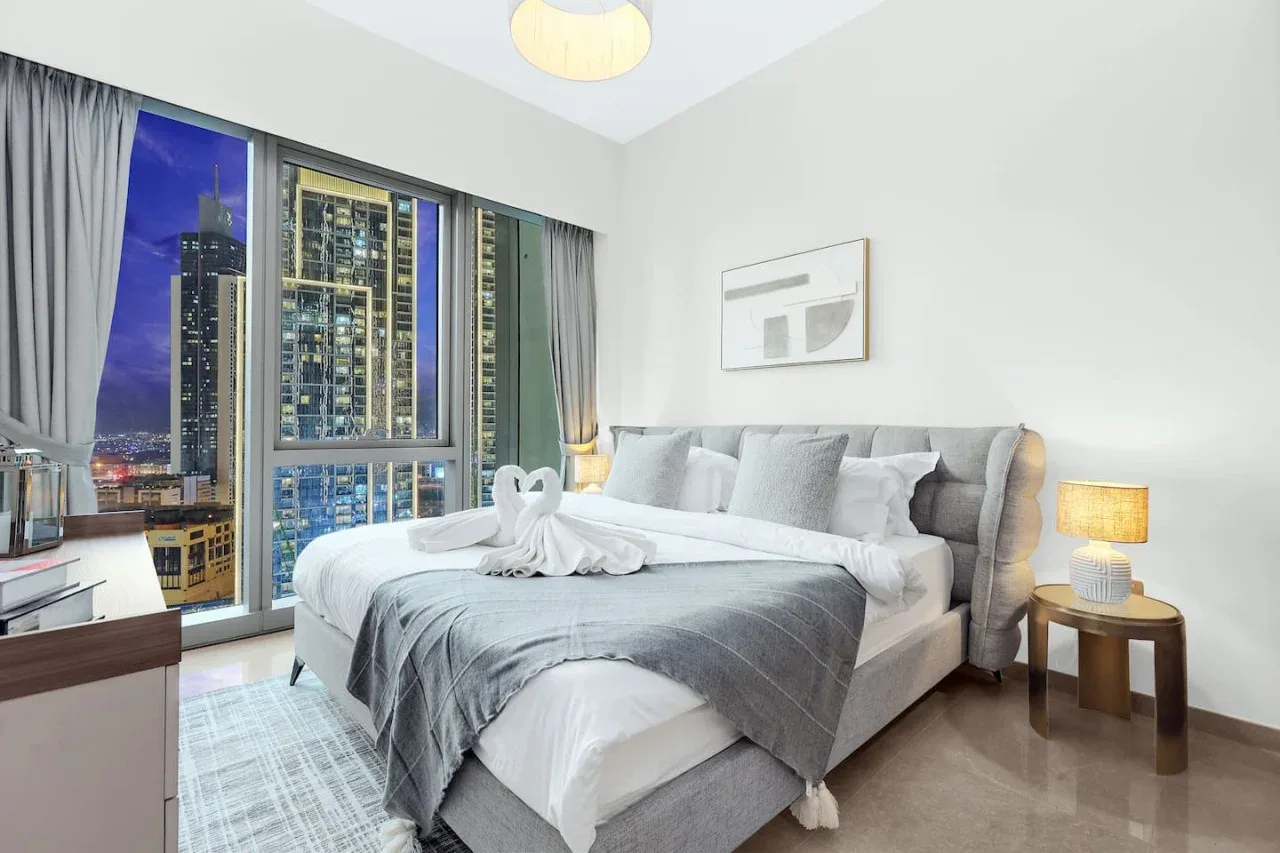 Un dormitorio con una cama grande junto a ventanales con vistas a edificios de la ciudad.