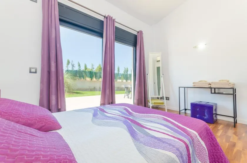 A bedroom with a bed dressed in purple bedding beside a large window.