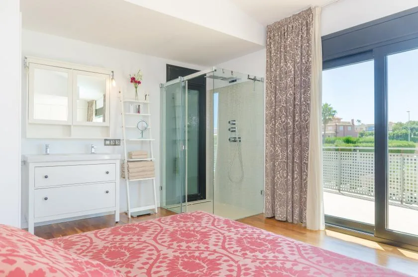 A bedroom with a patterned bedspread, vanity, and glass shower beside a balcony door.