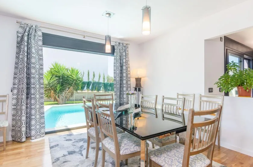 A bright dining area with a glass table beside large windows overlooking a pool.