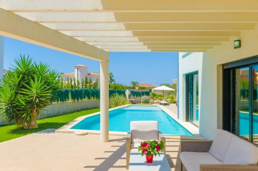 A covered patio with seating overlooks a swimming pool and landscaped garden.