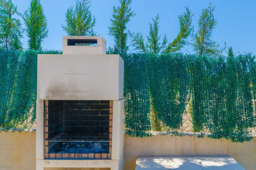 An outdoor barbecue grill stands beside a low wall and green privacy screen.