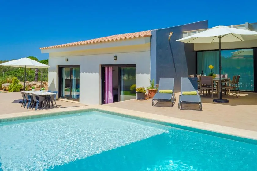 A sunny poolside patio beside a modern house, with loungers and outdoor dining.