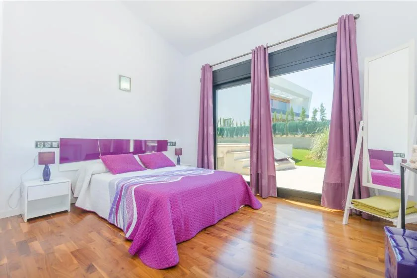 A bedroom with a bed dressed in pink bedding and curtains beside a large window.
