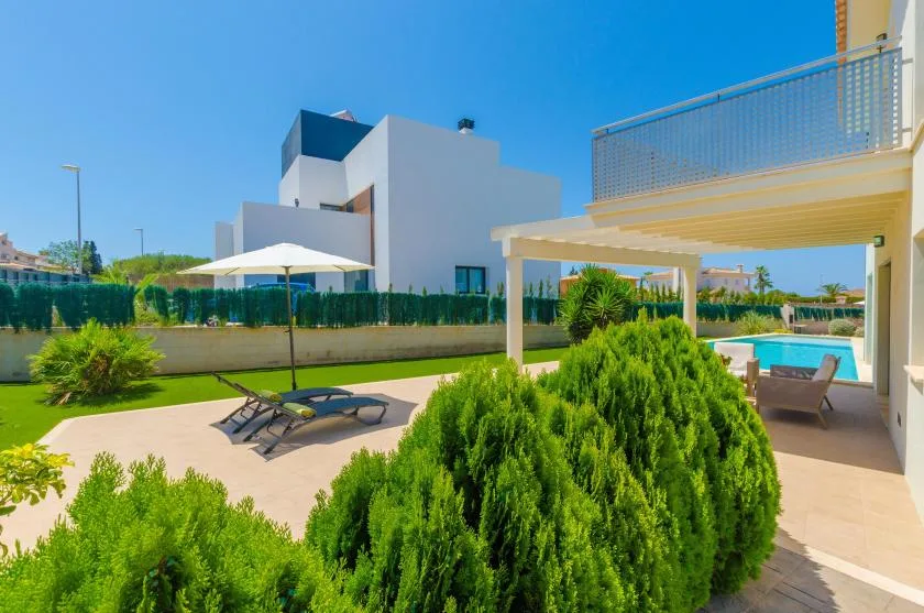 A sunny outdoor patio has lounge seating, greenery, and a swimming pool.