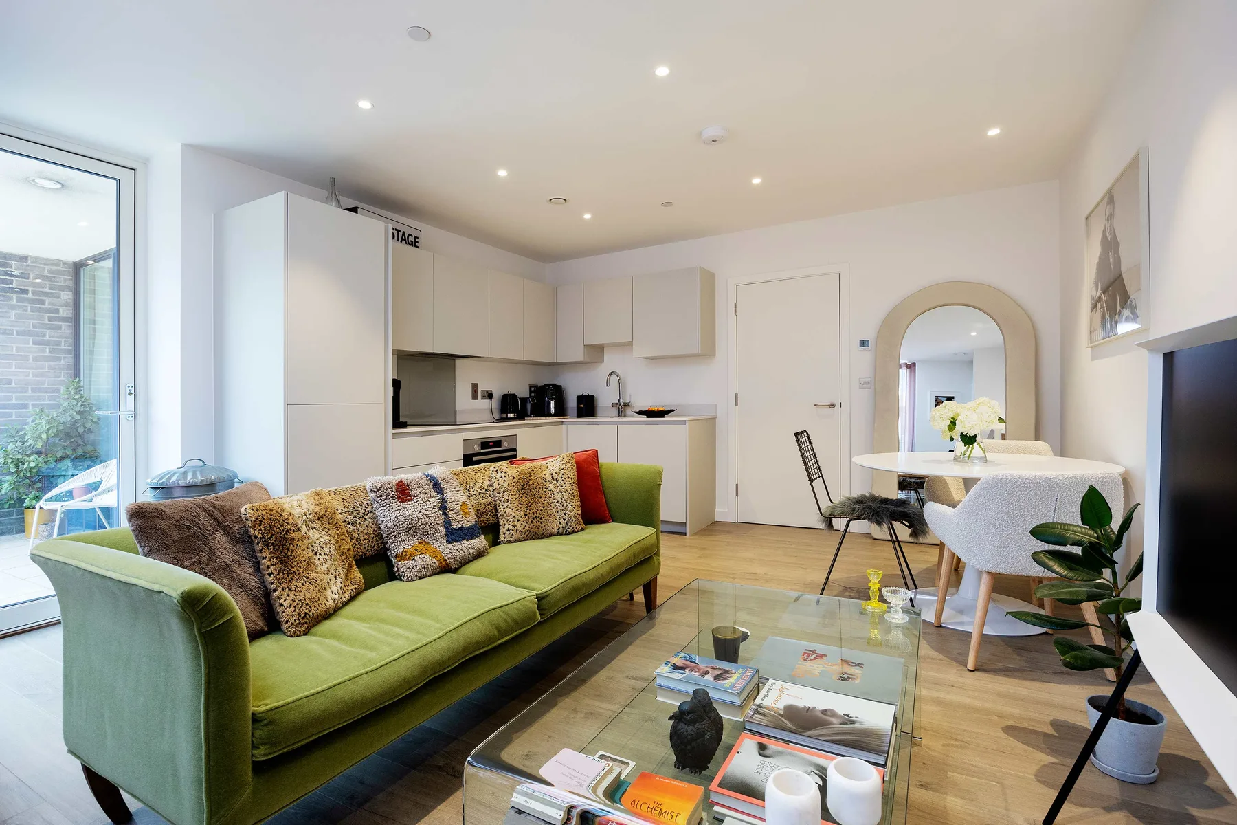 A bright living room with green seating, a glass coffee table, and an open kitchen.