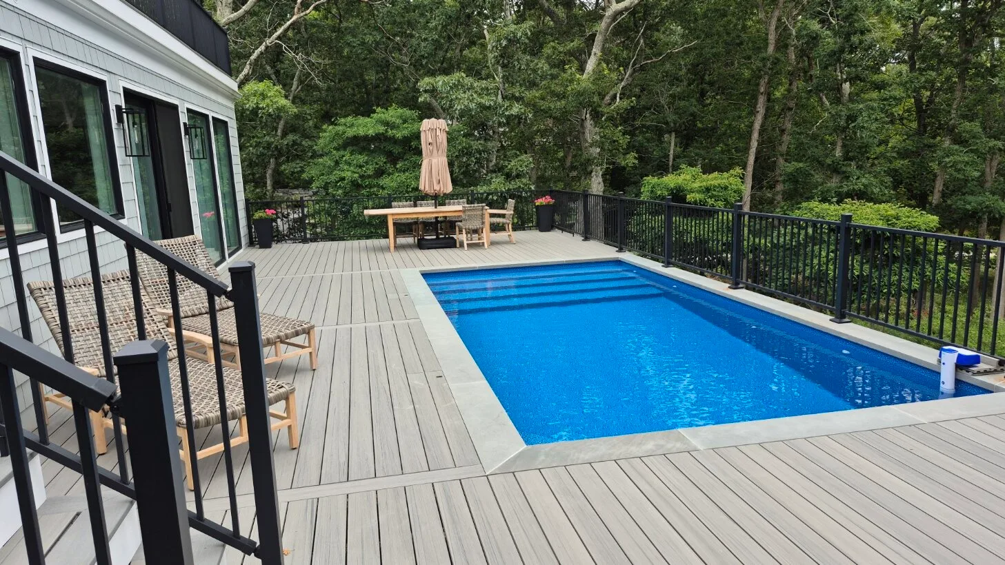 Una piscina al aire libre está junto a una amplia terraza y una cerca arbolada.