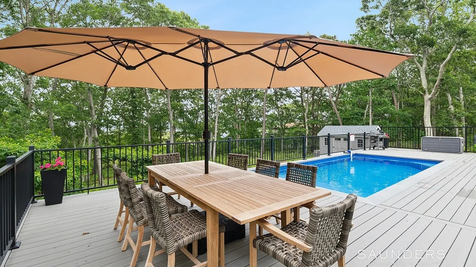 Una terraza exterior tiene una mesa larga bajo una gran sombrilla junto a una piscina.