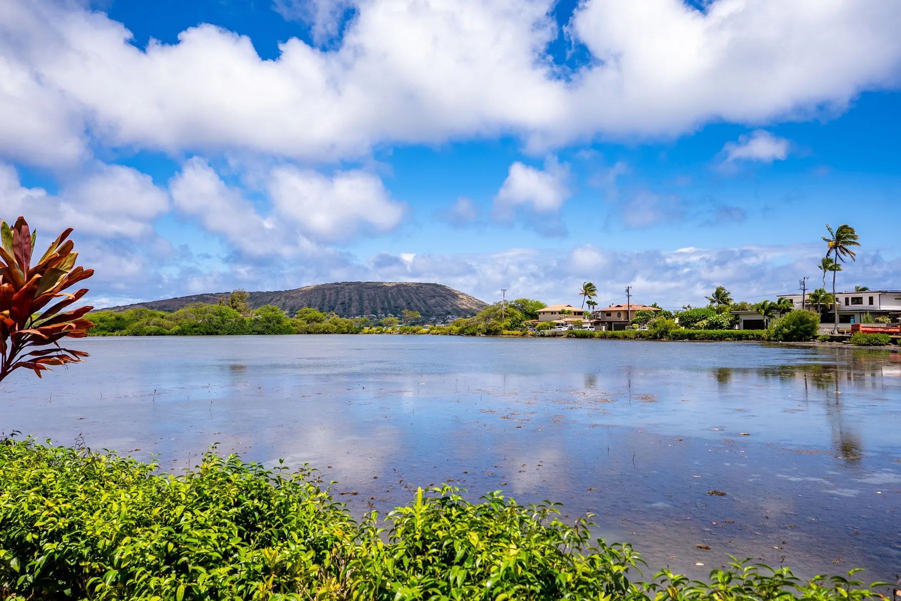 A calm lagoon reflects the cloudy sky, with greenery and homes along the shore.