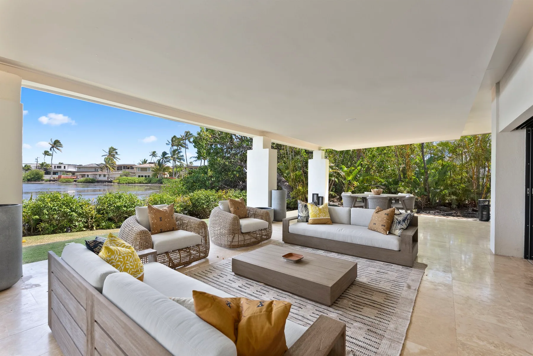 A covered outdoor lounge with cushioned seating and a low table overlooks a landscaped waterfront.