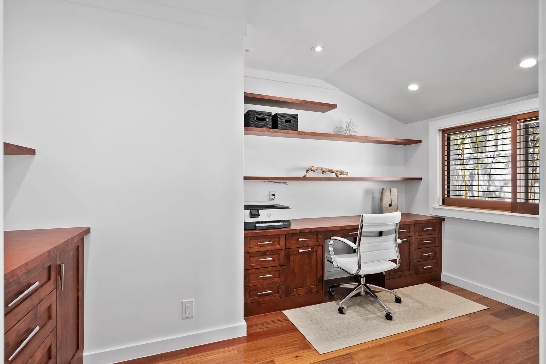 A home office with built-in wooden desks, open shelves, and a window.