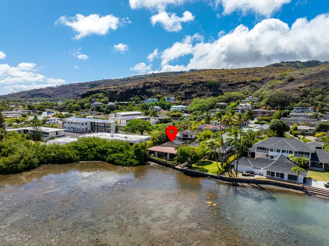 A coastal neighborhood of low-rise homes sits beside a calm inlet beneath a green hillside.