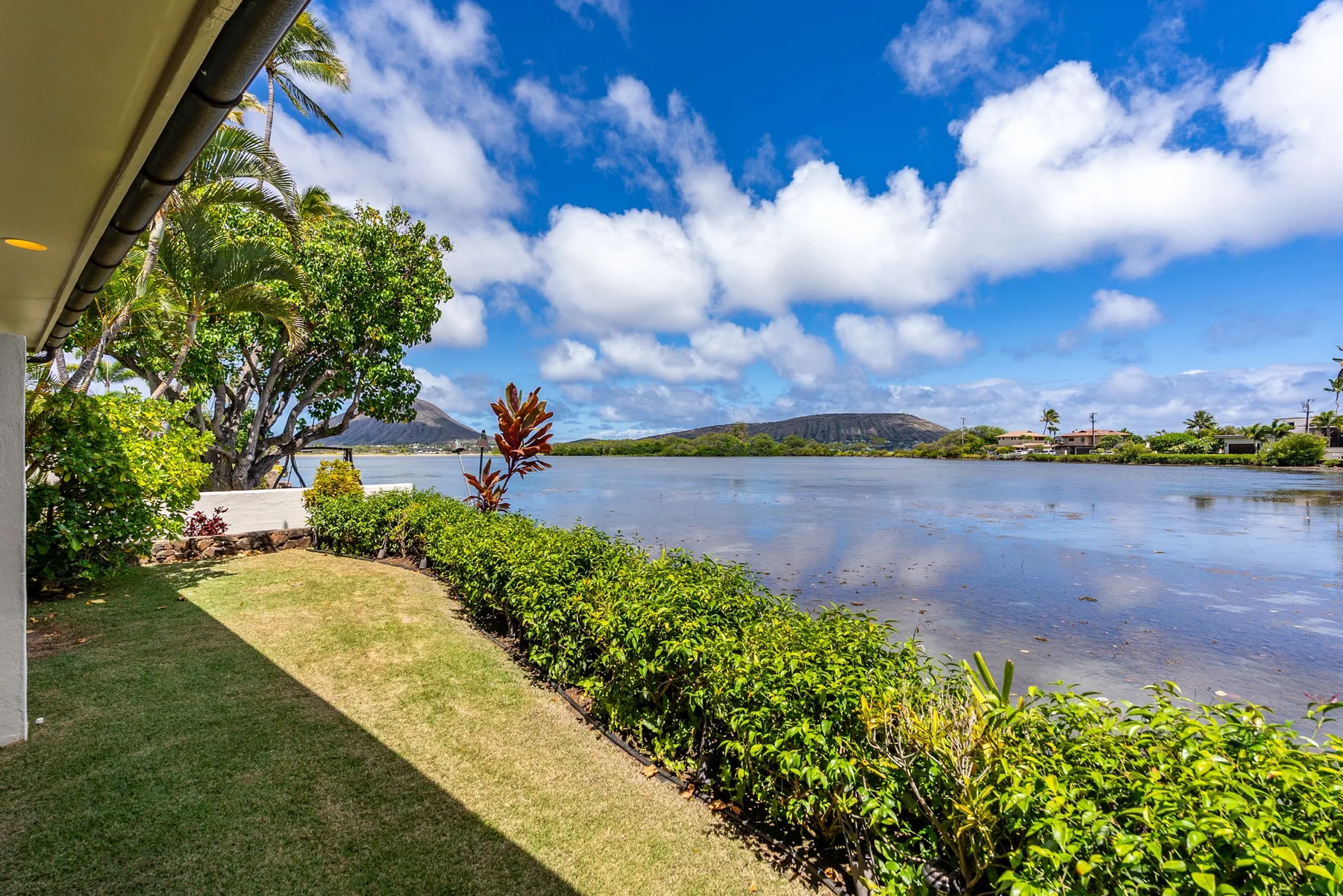 A landscaped lawn and hedge border a calm body of water beneath a cloudy blue sky.
