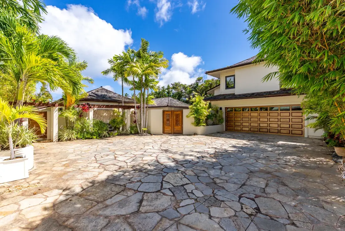 A house faces a broad stone-paved driveway, with a garage and tropical greenery.