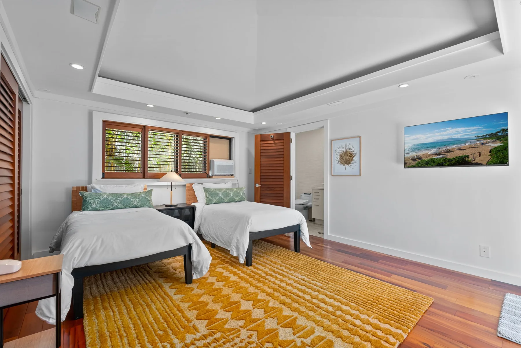 A bright bedroom with two beds, a large patterned rug, and windows with wooden blinds.