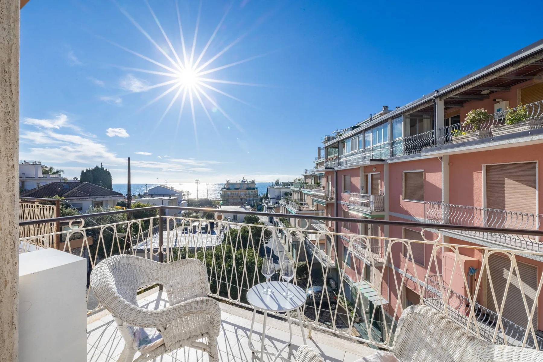 A sunny balcony with outdoor seating overlooks nearby buildings and the sea.