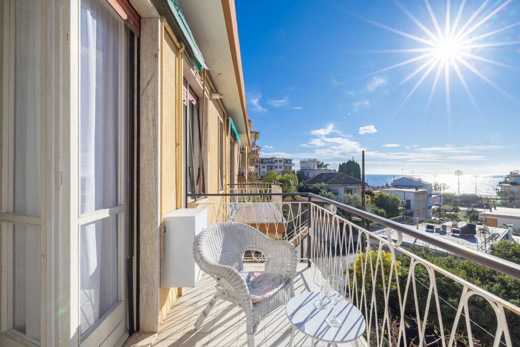A sunlit balcony with outdoor seating overlooks nearby buildings and the water.
