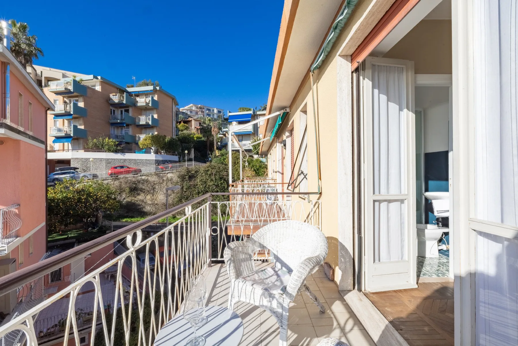 A sunlit balcony with white seating overlooks nearby apartment buildings.